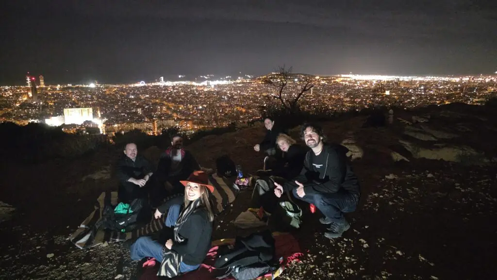 A group of people enjoying a nighttime picnic on a hill overlooking the illuminated cityscape of Barcelona, with blankets, snacks, and smiles under the dark sky.