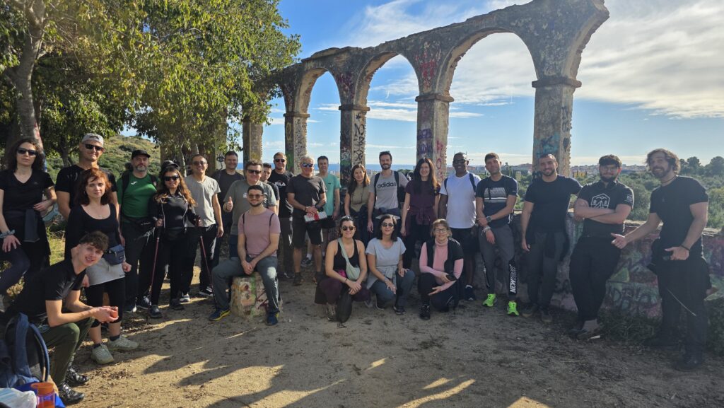 A group of international people gathered under stone arches at a scenic viewpoint in the Sierra de la Marina, with mountains and blue sky in the background